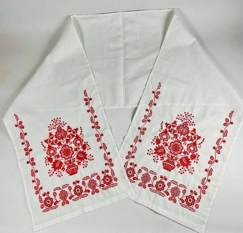 May include: White table runner with red embroidered floral designs. The design features two bouquets of flowers in a vase with leaves and stems.