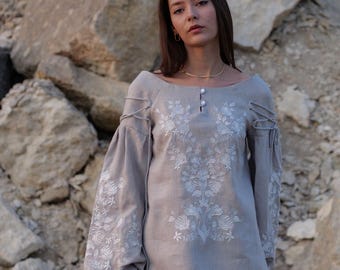 May include: A light gray linen blouse with white floral embroidery. The blouse features long, gathered sleeves and decorative rope detailing on the shoulders. It has a boat neckline and is fastened with three small buttons.