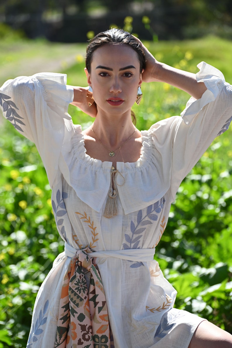 May include: A white linen dress with embroidered floral details, a tassel tie, and a colorful patterned sash. The dress has a ruffled neckline and a relaxed fit.