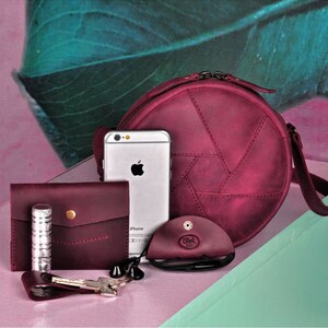 May include: A collection of burgundy leather accessories, including a round crossbody bag, wallet, cardholder, and eyeglass case. A silver iPhone, keys, and lipstick are also present. The bag has a unique stitched design.