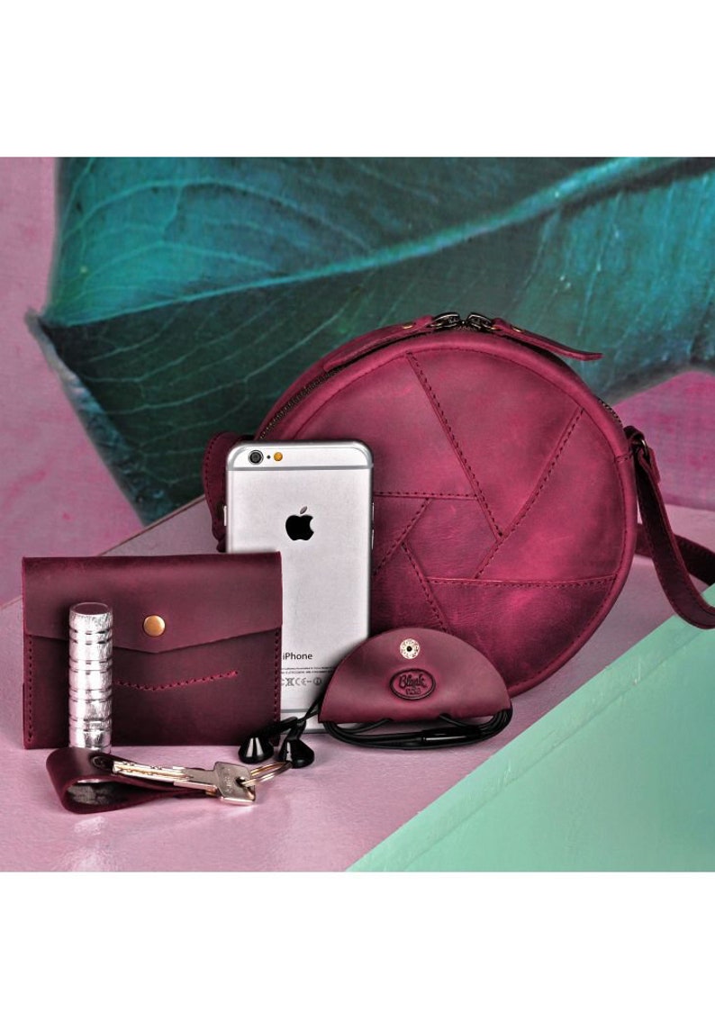May include: A collection of burgundy leather accessories, including a round crossbody bag, wallet, cardholder, and eyeglass case. A silver iPhone, keys, and lipstick are also present. The bag has a unique stitched design.