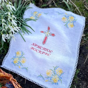 以下が含まれることがあります： 赤い十字架と黄色と青の花柄の刺繍が施された白いリネン布。布には繊細なレースのトリムがあり、赤い文字で「ХРИСТОС ВОСКРЕС」と書かれています。装飾されたイースターエッグのバスケットが前景にあります。