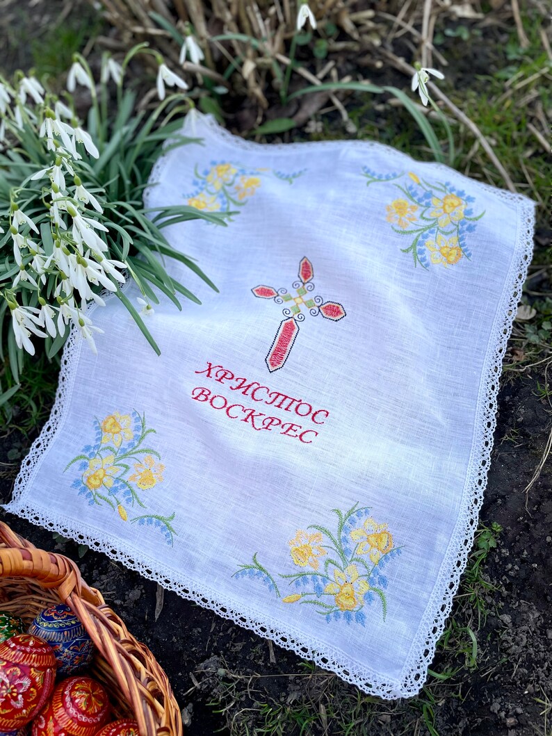 以下が含まれることがあります： 赤い十字架と黄色と青の花柄の刺繍が施された白いリネン布。布には繊細なレースのトリムがあり、赤い文字で「ХРИСТОС ВОСКРЕС」と書かれています。装飾されたイースターエッグのバスケットが前景にあります。