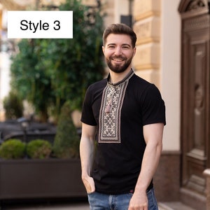Peut inclure: Un t-shirt noir &agrave; manches courtes avec un motif brod&eacute; marron sur le devant. Le t-shirt est port&eacute; par une personne avec une barbe et un sourire. La personne est debout devant un b&acirc;timent.