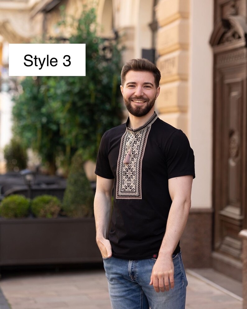 Peut inclure: Un t-shirt noir &agrave; manches courtes avec un motif brod&eacute; marron sur le devant. Le t-shirt est port&eacute; par une personne avec une barbe et un sourire. La personne est debout devant un b&acirc;timent.