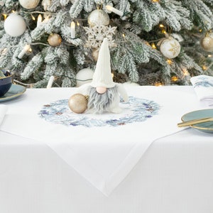 K&ouml;nnte beinhalten: Eine festliche Weihnachtstafel mit wei&szlig;er Tischdecke, blau-goldger&auml;nderten Teetassen, Tellern und Servietten. Eine Wichtelfigur und eine goldene Kugel liegen auf einem wei&szlig;en Tischl&auml;ufer mit blauem Blumendesign. Ein dekorierter Weihnachtsbaum befindet sich im Hintergrund.