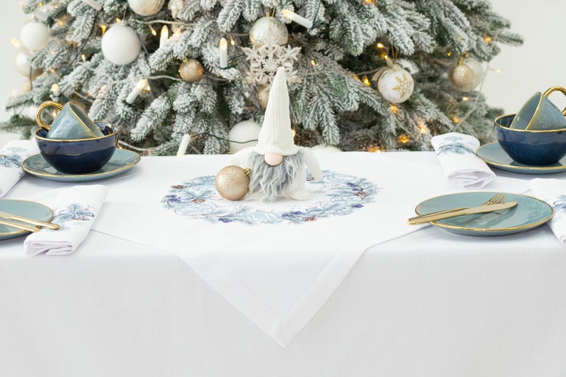 K&ouml;nnte beinhalten: Eine festliche Weihnachtstafel mit wei&szlig;er Tischdecke, blau-goldger&auml;nderten Teetassen, Tellern und Servietten. Eine Wichtelfigur und eine goldene Kugel liegen auf einem wei&szlig;en Tischl&auml;ufer mit blauem Blumendesign. Ein dekorierter Weihnachtsbaum befindet sich im Hintergrund.