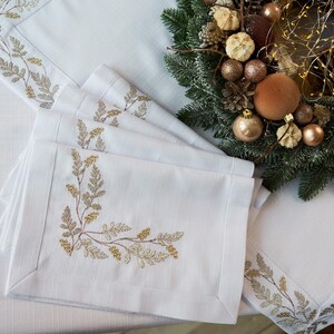 Puede incluir: Manteles individuales y servilletas de lino blanco con dise&ntilde;os bot&aacute;nicos dorados y beige bordados. Una corona navide&ntilde;a con adornos dorados y pi&ntilde;as est&aacute; en el fondo. Los manteles individuales y servilletas est&aacute;n cuidadosamente colocados sobre un mantel blanco.