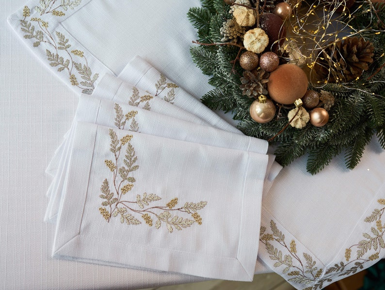 Puede incluir: Manteles individuales y servilletas de lino blanco con dise&ntilde;os bot&aacute;nicos dorados y beige bordados. Una corona navide&ntilde;a con adornos dorados y pi&ntilde;as est&aacute; en el fondo. Los manteles individuales y servilletas est&aacute;n cuidadosamente colocados sobre un mantel blanco.