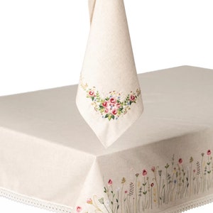 May include: Cream-colored linen tablecloth and matching napkin, both featuring embroidered floral designs. The tablecloth has a delicate lace trim. The napkin is hanging, showcasing a floral embroidery. The tablecloth is on a table.