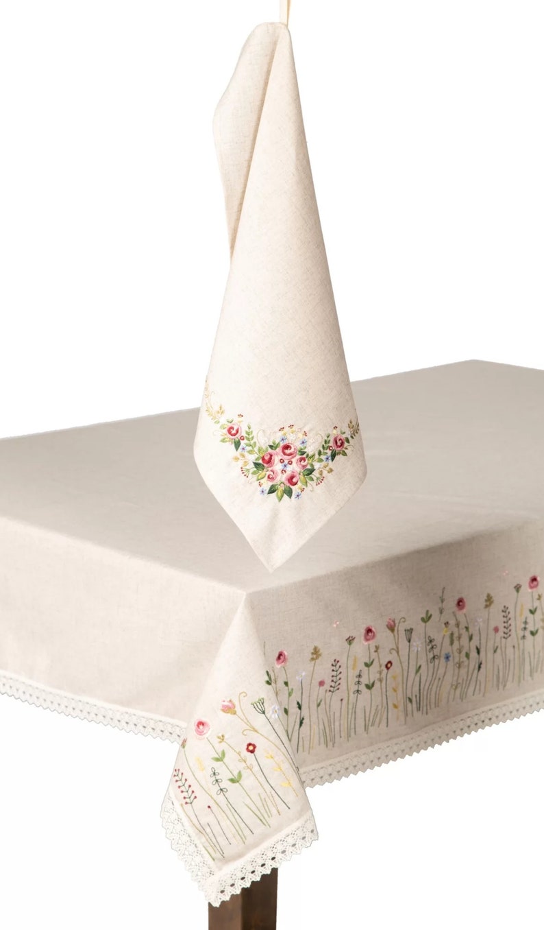 May include: Cream-colored linen tablecloth and matching napkin, both featuring embroidered floral designs. The tablecloth has a delicate lace trim. The napkin is hanging, showcasing a floral embroidery. The tablecloth is on a table.