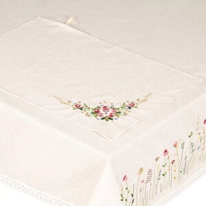 May include: Cream-colored linen tablecloth with embroidered floral details. A matching placemat is placed on the table. The edge of the tablecloth features a delicate floral border with red, pink, and yellow flowers.