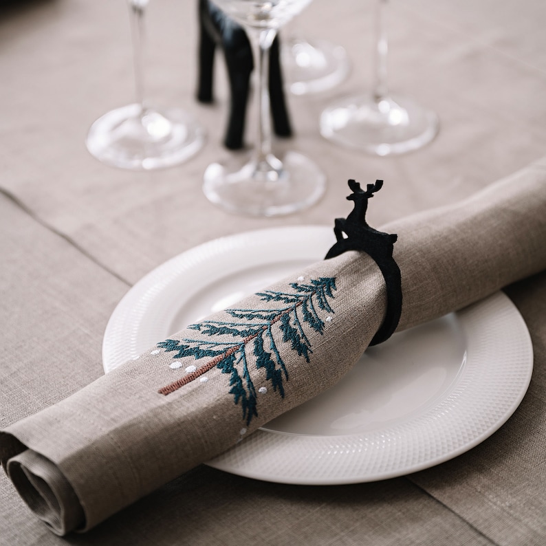 May include: A linen napkin with an embroidered Christmas tree design, rolled and held by a black reindeer napkin ring, rests on a white plate. The scene is set on a neutral-toned tablecloth, with wine glasses in the background.