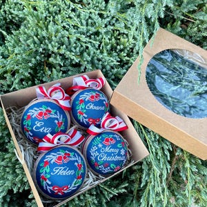 K&ouml;nnte beinhalten: Vier blaue und wei&szlig;e Weihnachtskugeln mit rot-wei&szlig;em Band. Die Kugeln sind mit gesticktem Stechpalmen, Glocken und Schneeflocken verziert. Die Kugeln sind mit den Namen "Eva", "Oksana", "Helen" und "Frohe Weihnachten" personalisiert.