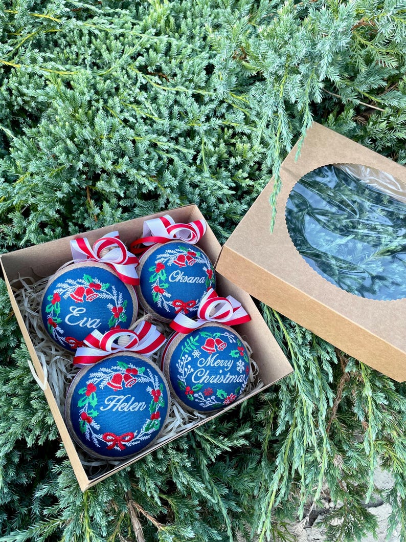 K&ouml;nnte beinhalten: Vier blaue und wei&szlig;e Weihnachtskugeln mit rot-wei&szlig;em Band. Die Kugeln sind mit gesticktem Stechpalmen, Glocken und Schneeflocken verziert. Die Kugeln sind mit den Namen "Eva", "Oksana", "Helen" und "Frohe Weihnachten" personalisiert.