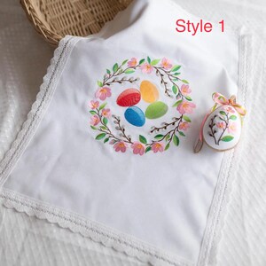 Op de afbeelding: Witte paaskleed met een bloemenkransborduurwerk met kleurrijke eieren. De doek heeft een kanten rand en de tekst "Style 1". Er is ook een paasei-ornament met bloemmotief en een roze lint aanwezig.