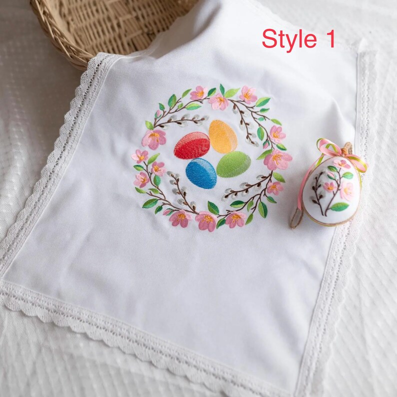 Op de afbeelding: Witte paaskleed met een bloemenkransborduurwerk met kleurrijke eieren. De doek heeft een kanten rand en de tekst "Style 1". Er is ook een paasei-ornament met bloemmotief en een roze lint aanwezig.
