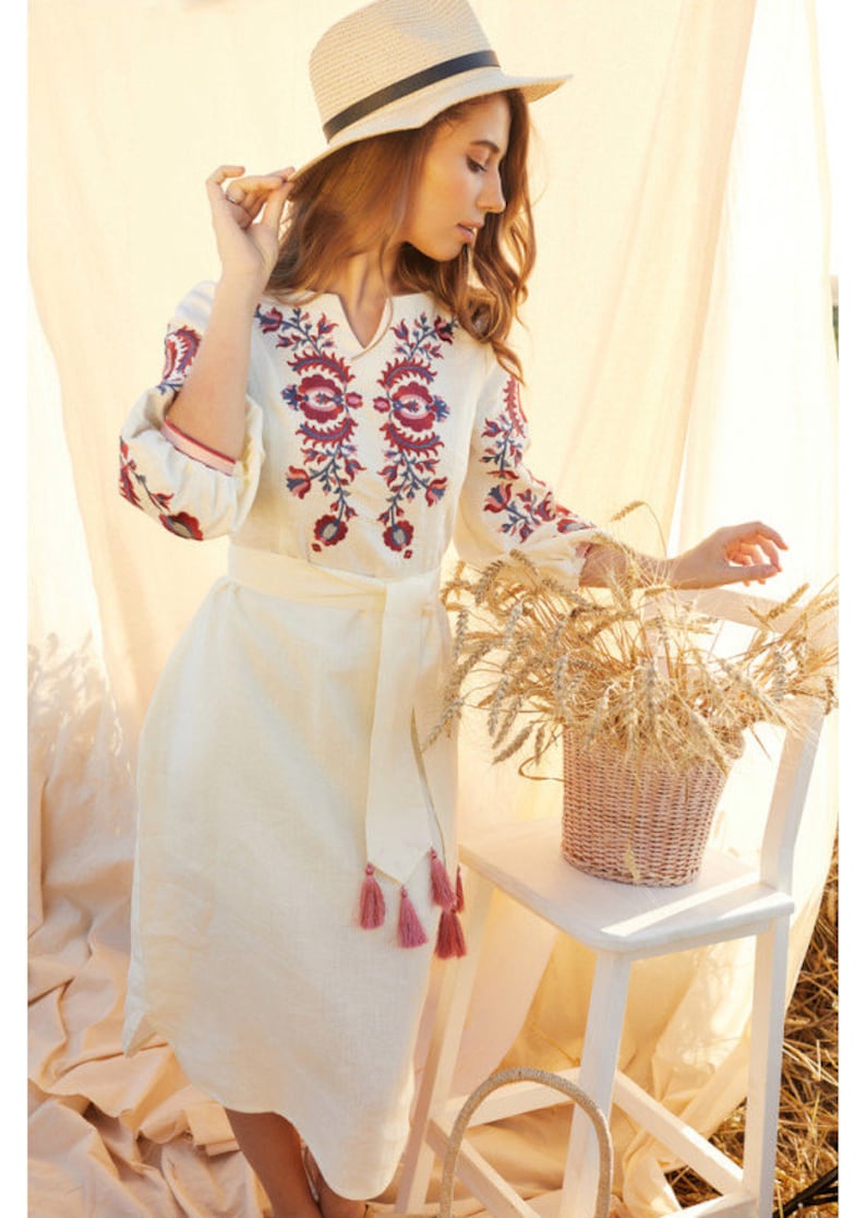 May include: A cream linen dress with red and blue floral embroidery. The dress has long sleeves, a V-neck, and a matching belt with pink tassels. A straw hat and a basket of wheat are also visible in the image.