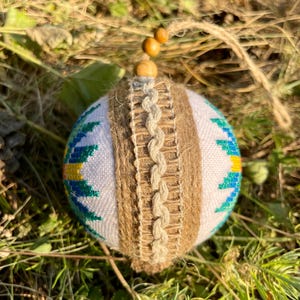 May include: A white Christmas ornament with a blue, green, and yellow geometric pattern. The ornament is wrapped in brown twine and has a wooden bead at the top.