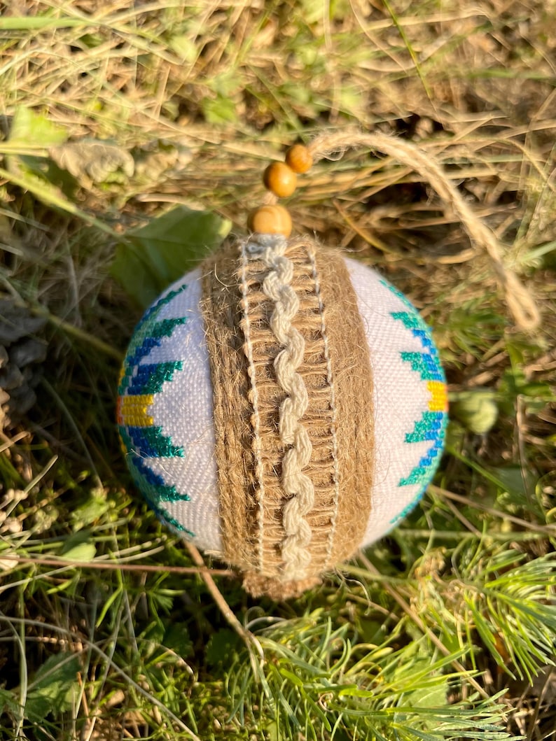 May include: A white Christmas ornament with a blue, green, and yellow geometric pattern. The ornament is wrapped in brown twine and has a wooden bead at the top.