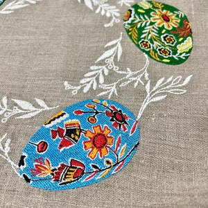以下が含まれることがあります： 白い花の刺繍が施されたリネン生地と、カラフルなイースターエッグ。卵は青、緑、赤、黄、黒の色の花柄で飾られています。