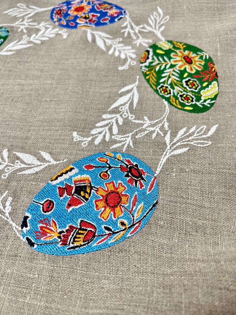 以下が含まれることがあります： 白い花の刺繍が施されたリネン生地と、カラフルなイースターエッグ。卵は青、緑、赤、黄、黒の色の花柄で飾られています。