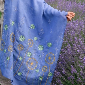 Pode incluir: Um xale azul claro com desenhos de dente-de-le&atilde;o bordados em tons de verde e amarelo. O xale est&aacute; drapeado sobre uma pessoa em p&eacute; num campo de lavanda.