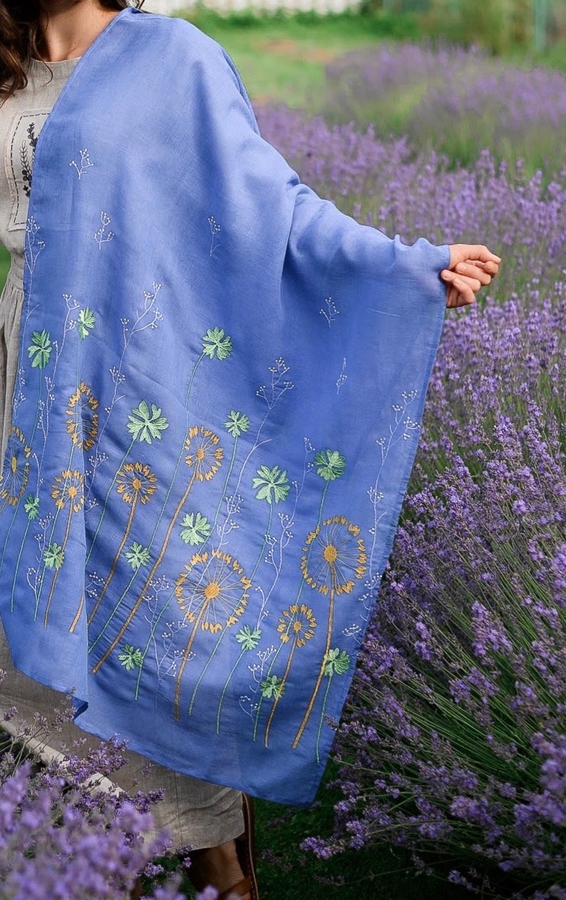 Pode incluir: Um xale azul claro com desenhos de dente-de-le&atilde;o bordados em tons de verde e amarelo. O xale est&aacute; drapeado sobre uma pessoa em p&eacute; num campo de lavanda.