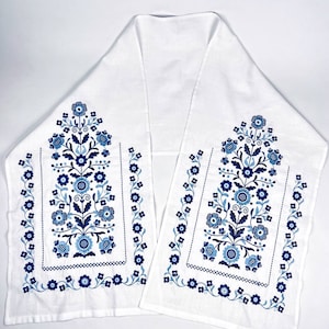 May include: White table runner with a floral design in shades of blue. The embroidered pattern features stylized flowers, leaves, and dotted accents. The runner is rectangular with a folded, symmetrical design.