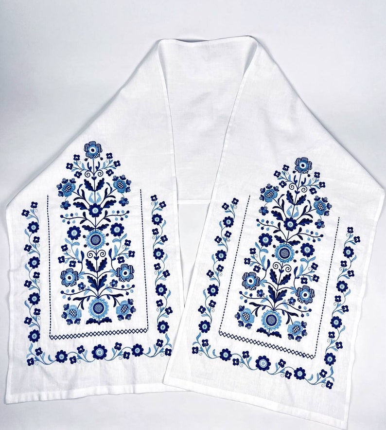 May include: White table runner with a floral design in shades of blue. The embroidered pattern features stylized flowers, leaves, and dotted accents. The runner is rectangular with a folded, symmetrical design.