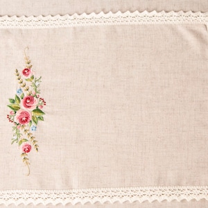 May include: Rectangular beige linen placemat with a floral embroidery design featuring pink roses, green leaves, and blue accents. The edges are trimmed with a white, scalloped lace.