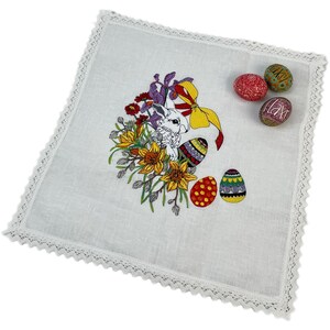 May include: White linen placemat with a lace border featuring an embroidered Easter bunny surrounded by colorful flowers and eggs.