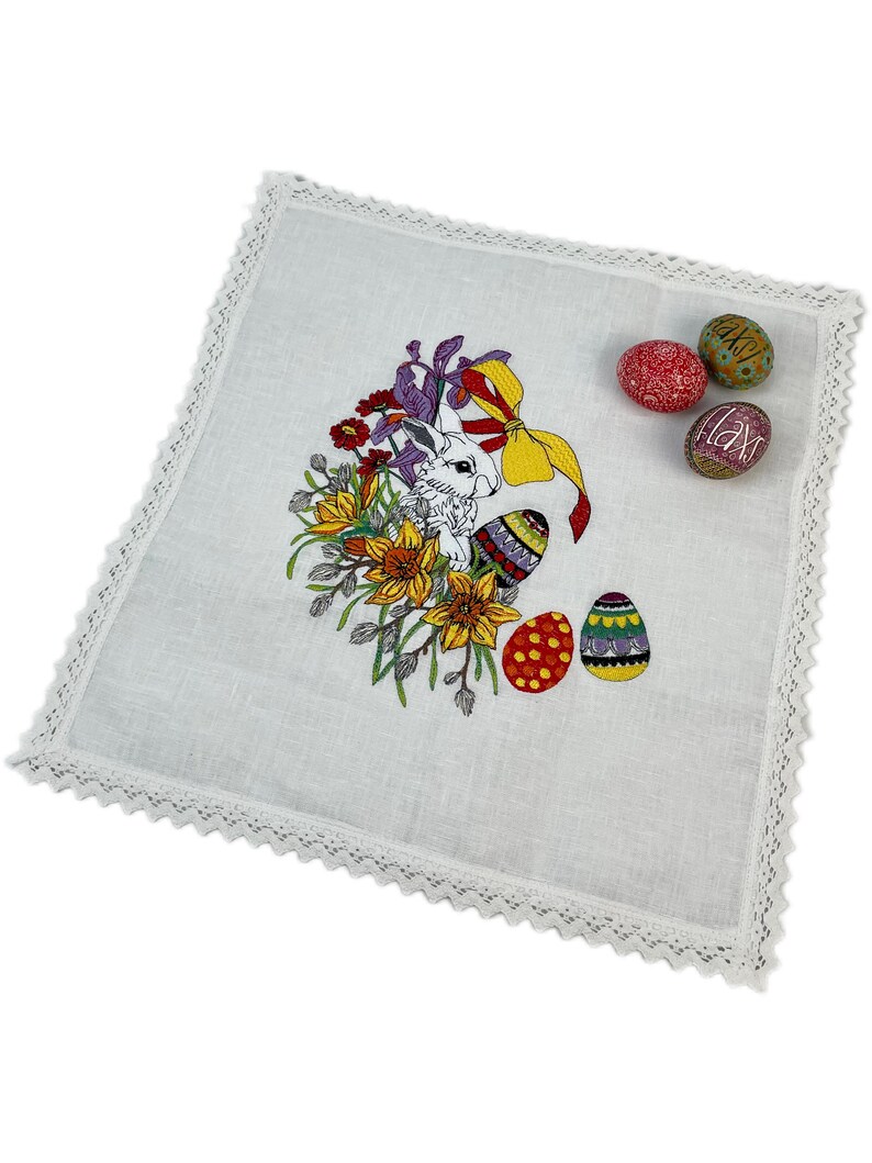 May include: White linen placemat with a lace border featuring an embroidered Easter bunny surrounded by colorful flowers and eggs.