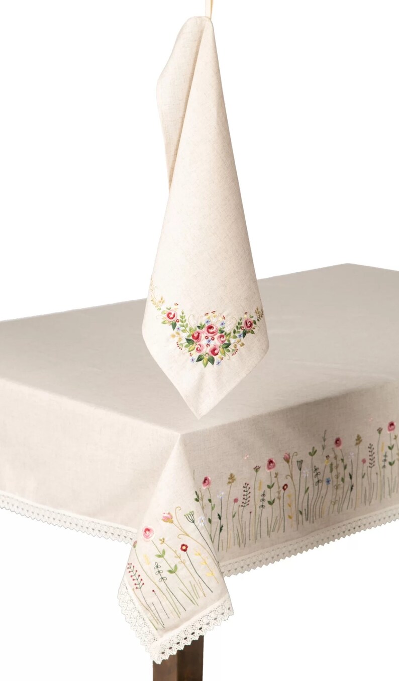 May include: Cream-colored linen tablecloth and matching napkin, both featuring embroidered floral designs in pink, red, and green. The tablecloth has a delicate lace trim along the edges. The napkin is hanging from a cream-colored ribbon.