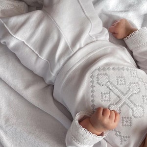May include: White baby clothing set with a cross embroidered on the front. The set includes a long-sleeved shirt and pants.