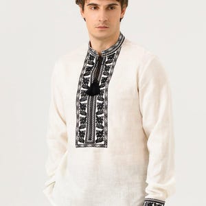 May include: Off-white long-sleeved shirt with intricate black floral embroidery on the collar, placket, and cuffs. The shirt features a mandarin collar and a black tassel detail. The fabric appears to be linen or a similar natural material.