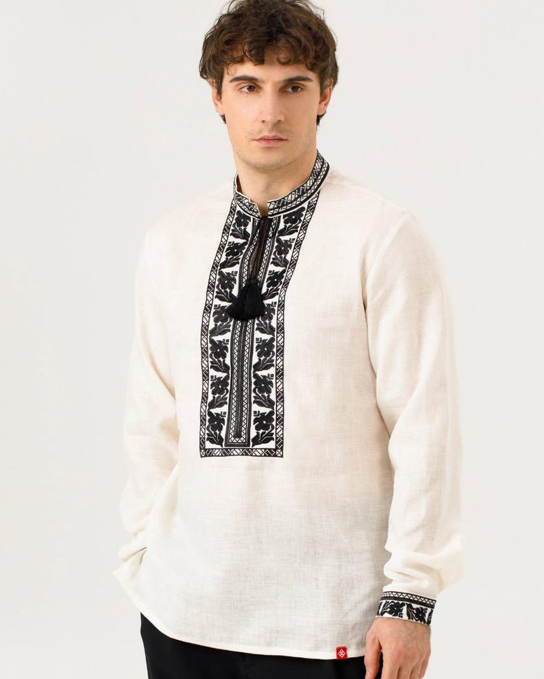 May include: Off-white long-sleeved shirt with intricate black floral embroidery on the collar, placket, and cuffs. The shirt features a mandarin collar and a black tassel detail. The fabric appears to be linen or a similar natural material.