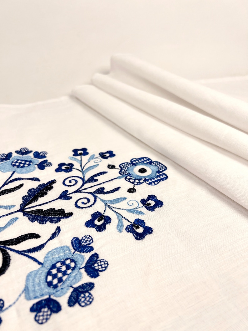 May include: White linen tablecloth with blue and black embroidered floral design. The embroidery features stylized flowers and leaves.