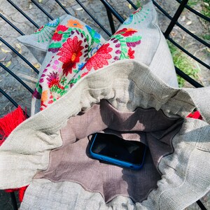 May include: A gray linen tote bag with a floral embroidered design. The bag is open and a blue smartphone is inside.