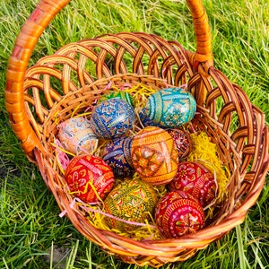 K&ouml;nnte beinhalten: Ein Weidenkorb gef&uuml;llt mit farbenfroh dekorierten Ostereiern. Die Eier weisen komplizierte Muster in Rot, Blau, Gelb und Gr&uuml;n auf. Der Korb liegt auf gelbem Stroh, umgeben von gr&uuml;nem Gras.