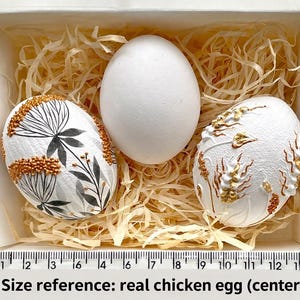 May include: Three decorated eggs in a white box with straw. Two eggs feature floral patterns in gold, black, and white. The central egg is a plain white chicken egg. Text on the image reads "Size reference: real chicken egg (center)".