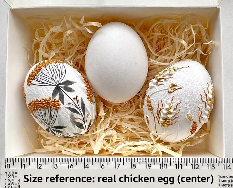 May include: Three decorated eggs in a white box with straw. Two eggs feature floral patterns in gold, black, and white. The central egg is a plain white chicken egg. Text on the image reads "Size reference: real chicken egg (center)".