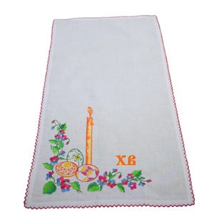 K&ouml;nnte beinhalten: Wei&szlig;es besticktes Leinen-Geschirrtuch mit einer gelben Kerze, drei bunten Ostereiern und gr&uuml;nen und rosa Blumen. Der Text "XB" ist in Orange gestickt.