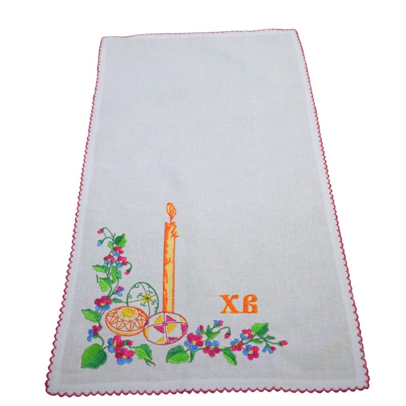 K&ouml;nnte beinhalten: Wei&szlig;es besticktes Leinen-Geschirrtuch mit einer gelben Kerze, drei bunten Ostereiern und gr&uuml;nen und rosa Blumen. Der Text "XB" ist in Orange gestickt.