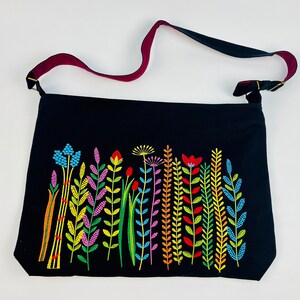 May include: Black tote bag with a colorful embroidered floral design. The bag features a long strap with a burgundy underside. The embroidered flowers are in shades of blue, yellow, red, and purple.