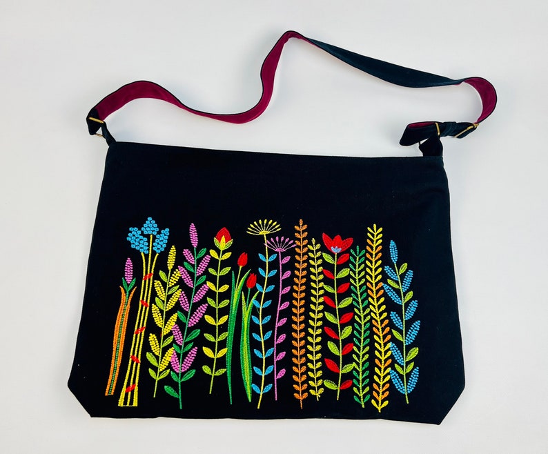 May include: Black tote bag with a colorful embroidered floral design. The bag features a long strap with a burgundy underside. The embroidered flowers are in shades of blue, yellow, red, and purple.