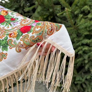 May include: A white shawl with a floral and paisley pattern and a fringe border. The shawl is held in a hand with red nail polish.