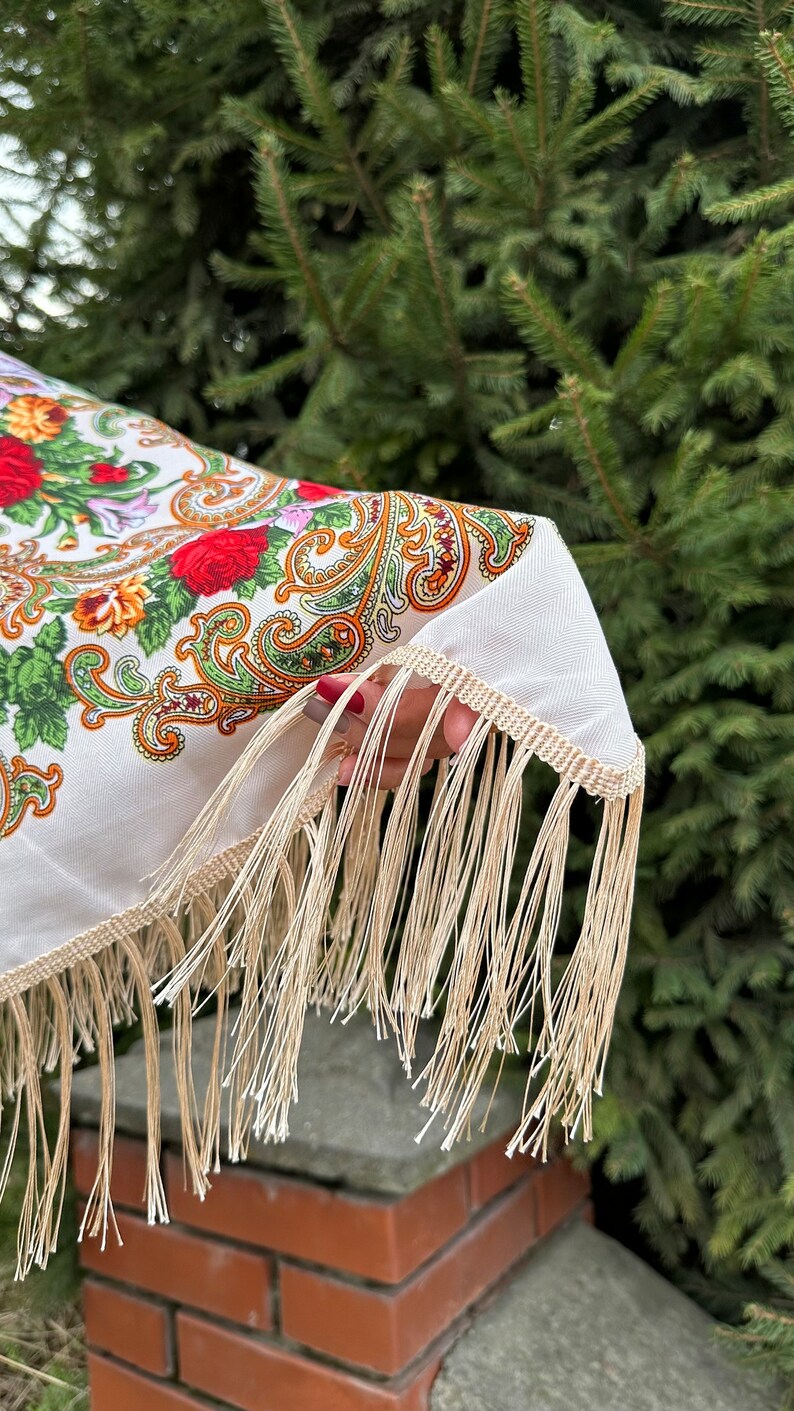 May include: A white shawl with a floral and paisley pattern and a fringe border. The shawl is held in a hand with red nail polish.