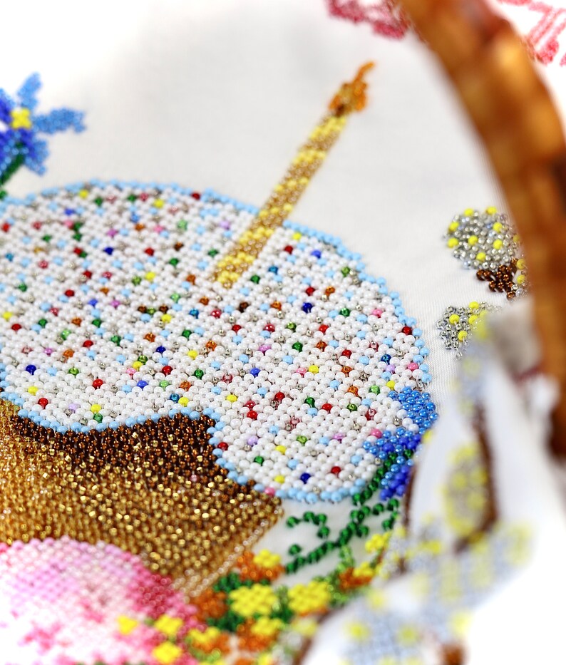 May include: Close-up of a white fabric with a beaded design of a cupcake with a lit candle. The cupcake has a brown base and white frosting with colourful sprinkles. Blue and yellow flowers are also beaded.