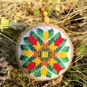 May include: A round, white ornament with a green, red, yellow, and orange geometric pattern embroidered on it. The ornament is made of fabric and has a burlap backing. It is hanging from a wooden bead.